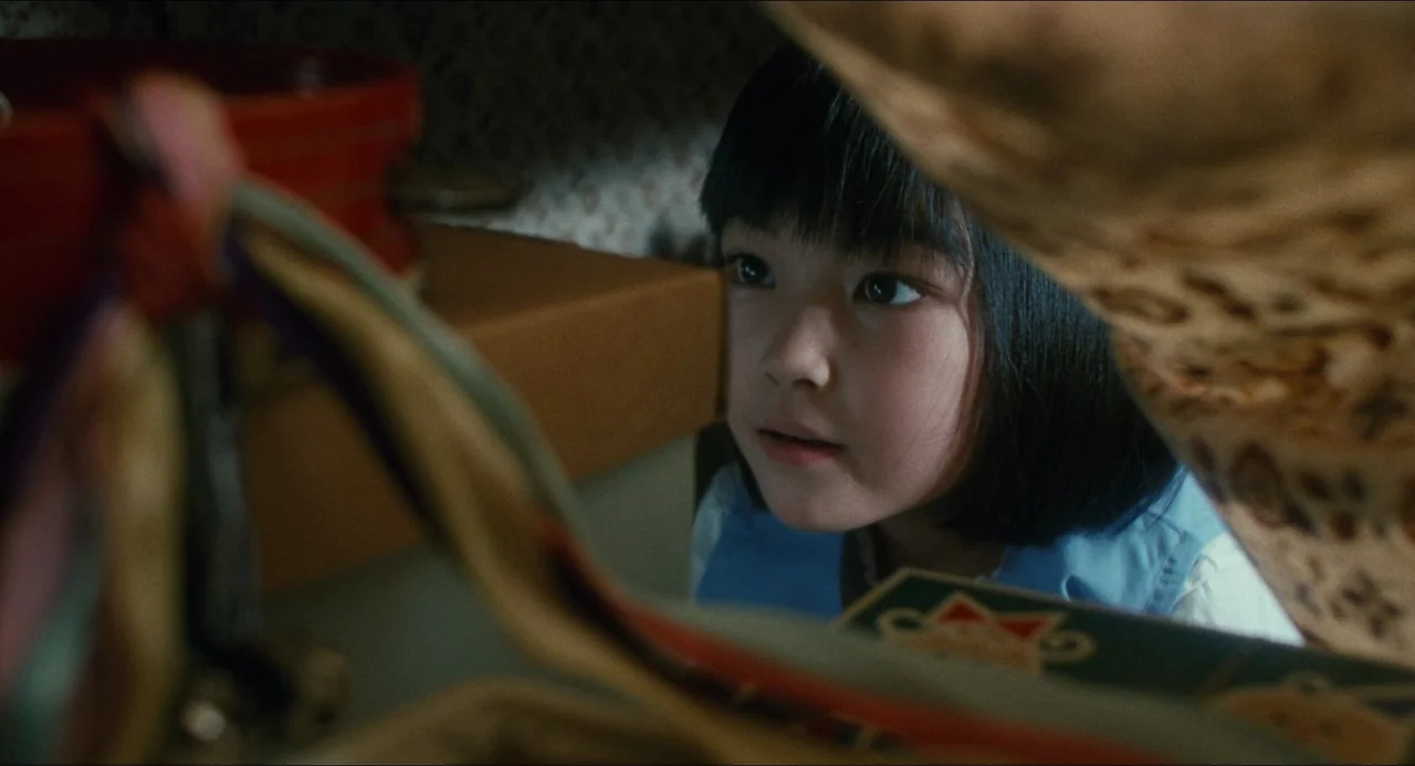 Dark Water (2002) movie still: closeup — A child peers through an opening between stacked household items in a dimly lit area. The space contains…
