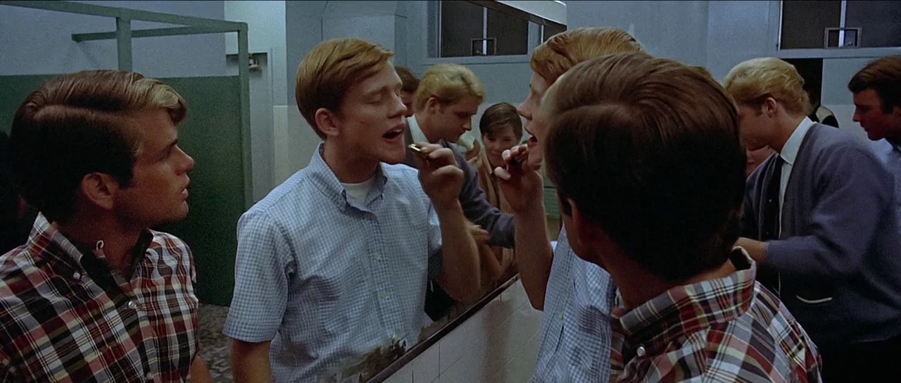 American Graffiti (1973) movie still: medium — A group of young men stand in a school restroom with white tiled walls. One young man is applying…