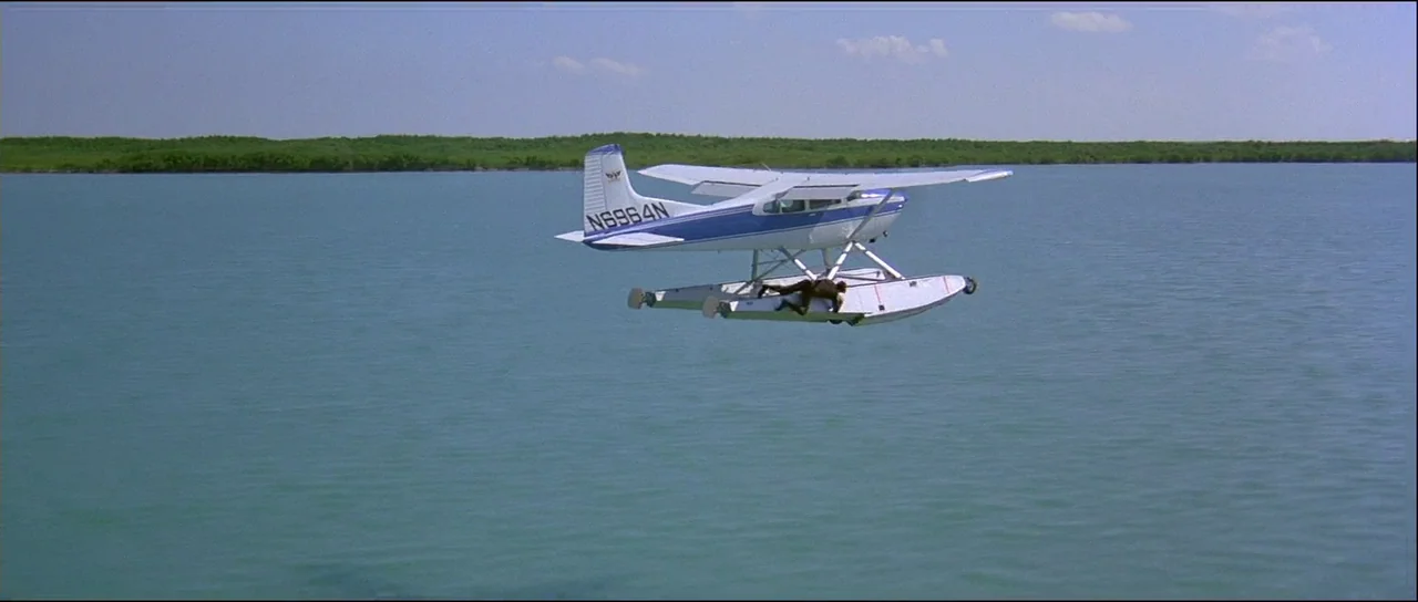 Licence to Kill (1989) movie still: wide — A white and blue seaplane flies low over calm, turquoise open water. A person lies prone on top of the…