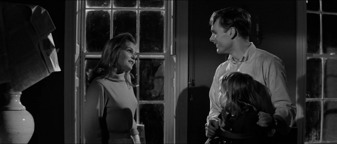Bunny Lake Is Missing (1965) movie still: medium — A dimly lit room with large windows showing night beyond the glass. A woman stands opposite a man…
