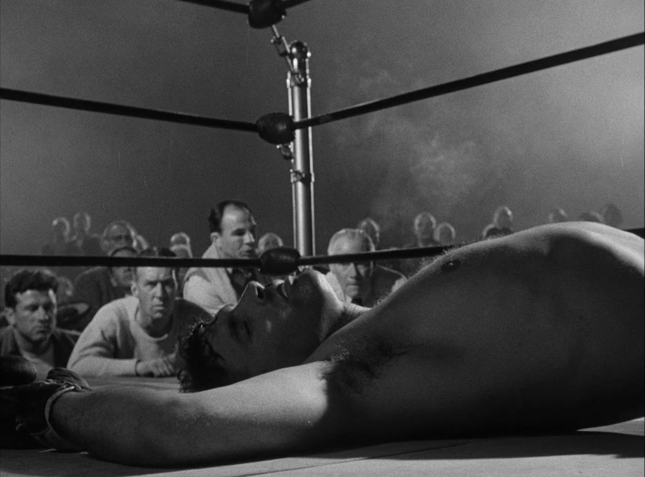 The Killers (1946) movie still: medium — A boxing ring canvas viewed from a low angle with spectators visible behind ropes. A male boxer lies…
