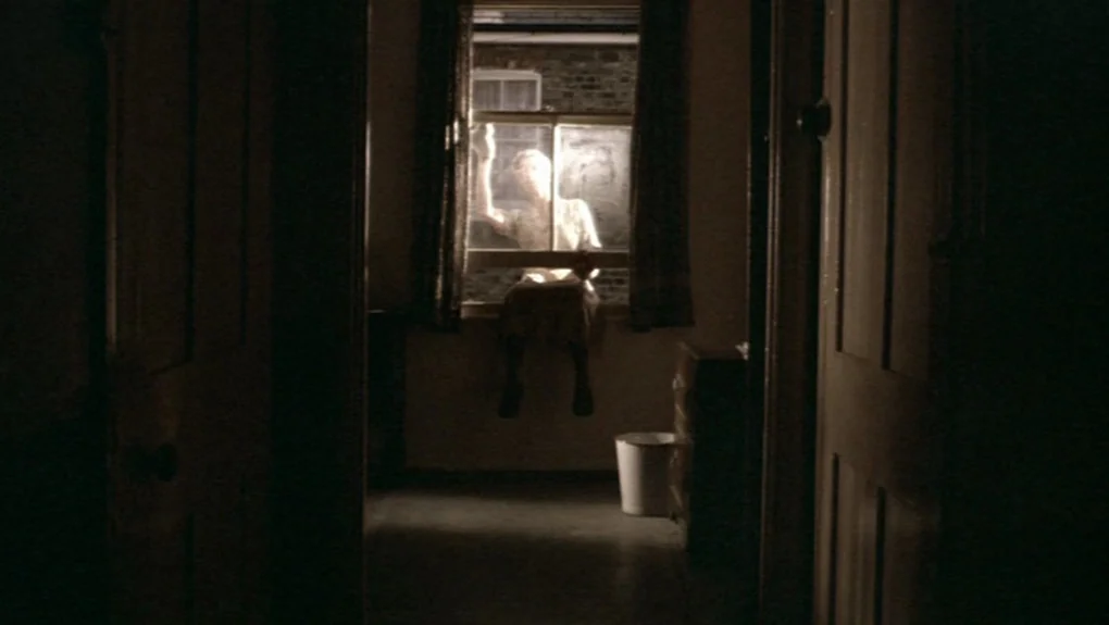 Distant Voices, Still Lives (1988) movie still: medium — A dark, sparsely furnished room is framed between two wooden door frames. A woman sits…