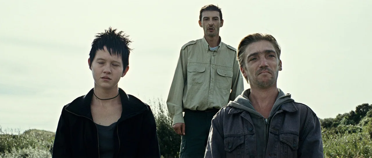 Outside Satan (2011) movie still: medium — Three individuals stand close together in an overgrown field under a bright overcast sky. A woman with…