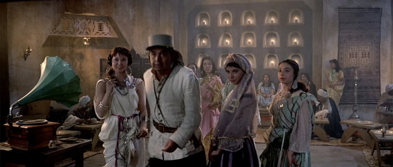 Legend of the Lost (1957) movie still: medium — A room with textured walls featuring numerous recessed alcoves holding small lit candles. A man and…