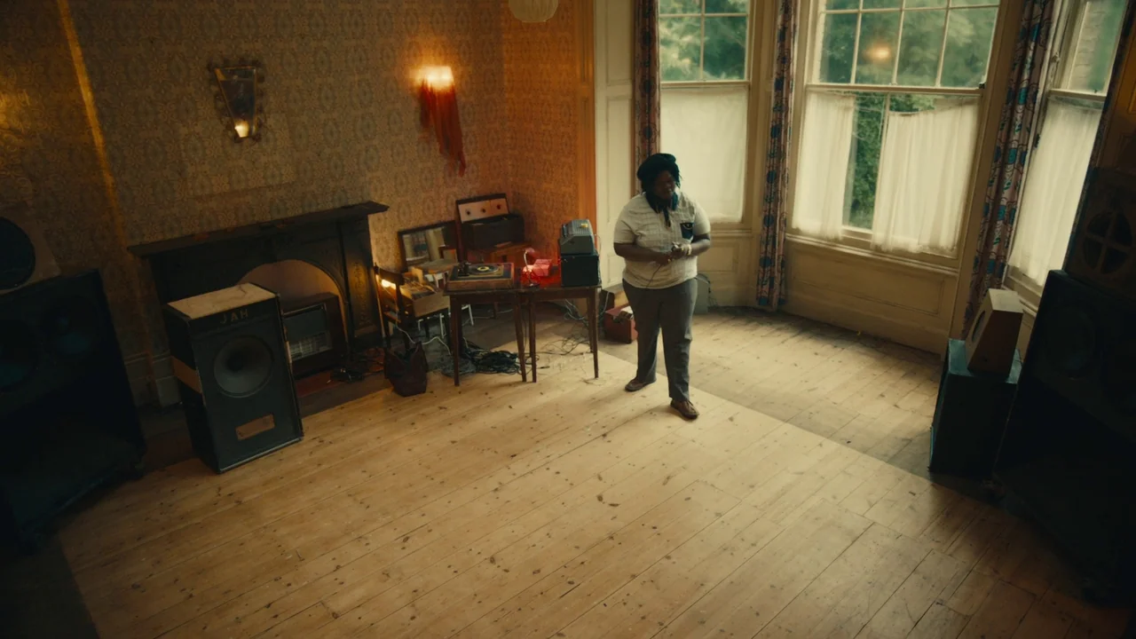 Lover’s Rock (2020) movie still: fullbody — A spacious room features patterned vintage wallpaper, wood floors, and large bay windows. A man stands…