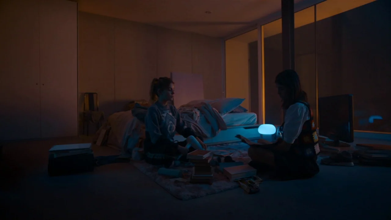 Amanda (2022) movie still: wide — A modern bedroom with floor-to-ceiling glass walls is dimly lit at night. Two young women sit on a shag rug…