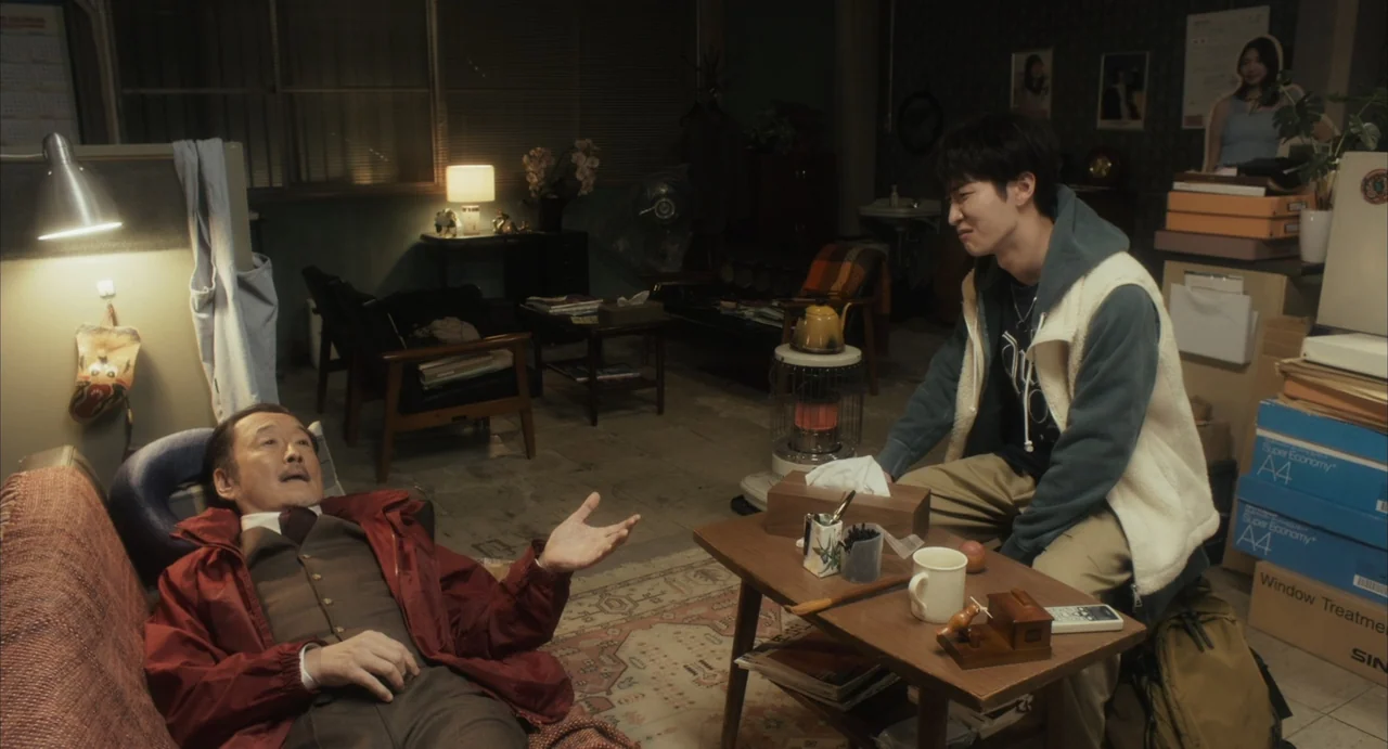 Stigmatized Properties: Possession (2025) movie still: medium — A cluttered apartment interior features various stacked boxes, a desk lamp, and home…