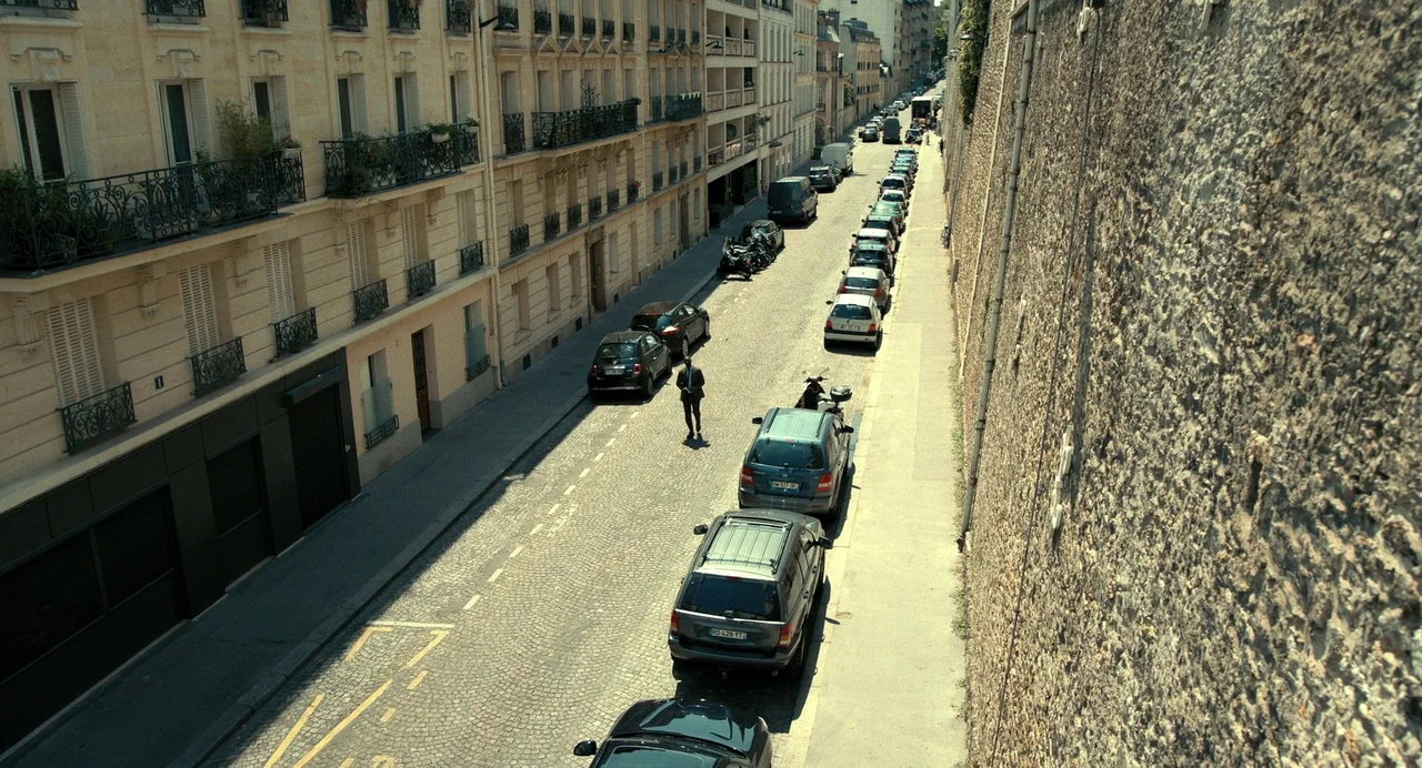 The Killer (2024) (2024) movie still: wide — A narrow cobblestone Parisian road is flanked by a tall stone wall and classic apartments. A lone man…