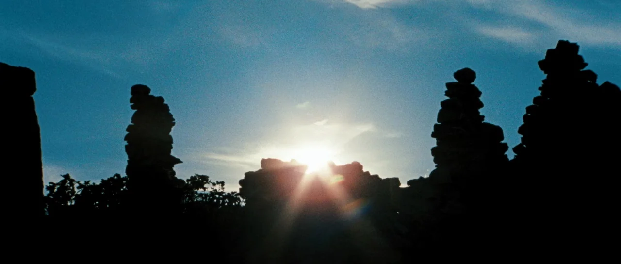 Let The Corpses Tan (2017) movie still: wide — Several stacks of balanced stones stand as dark silhouettes against a bright blue sky. The sun is…