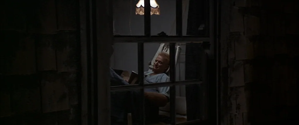 Popeye (1980) movie still: medium — A dark, dimly lit room is visible through an exterior window pane at night. A man sits in a chair under a…
