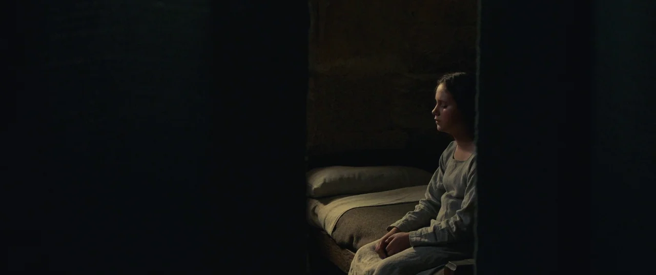 Benedetta (2021) movie still: medium — A stone cell contains a narrow wooden bed with a single pillow. A girl sits at the edge of the bed in…