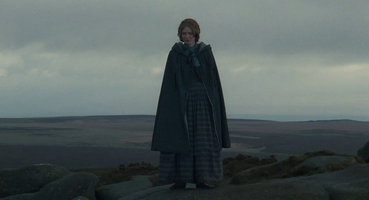 Jane Eyre (2011) movie still: fullbody — A vast, desolate moorland under a heavy, grey overcast sky with sparse vegetation. A young woman in period…