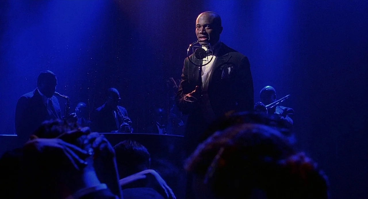 Chicago (2002) movie still: medium — A dimly lit nightclub features a man singing before a live brass band. The stage is bathed in deep blue light…