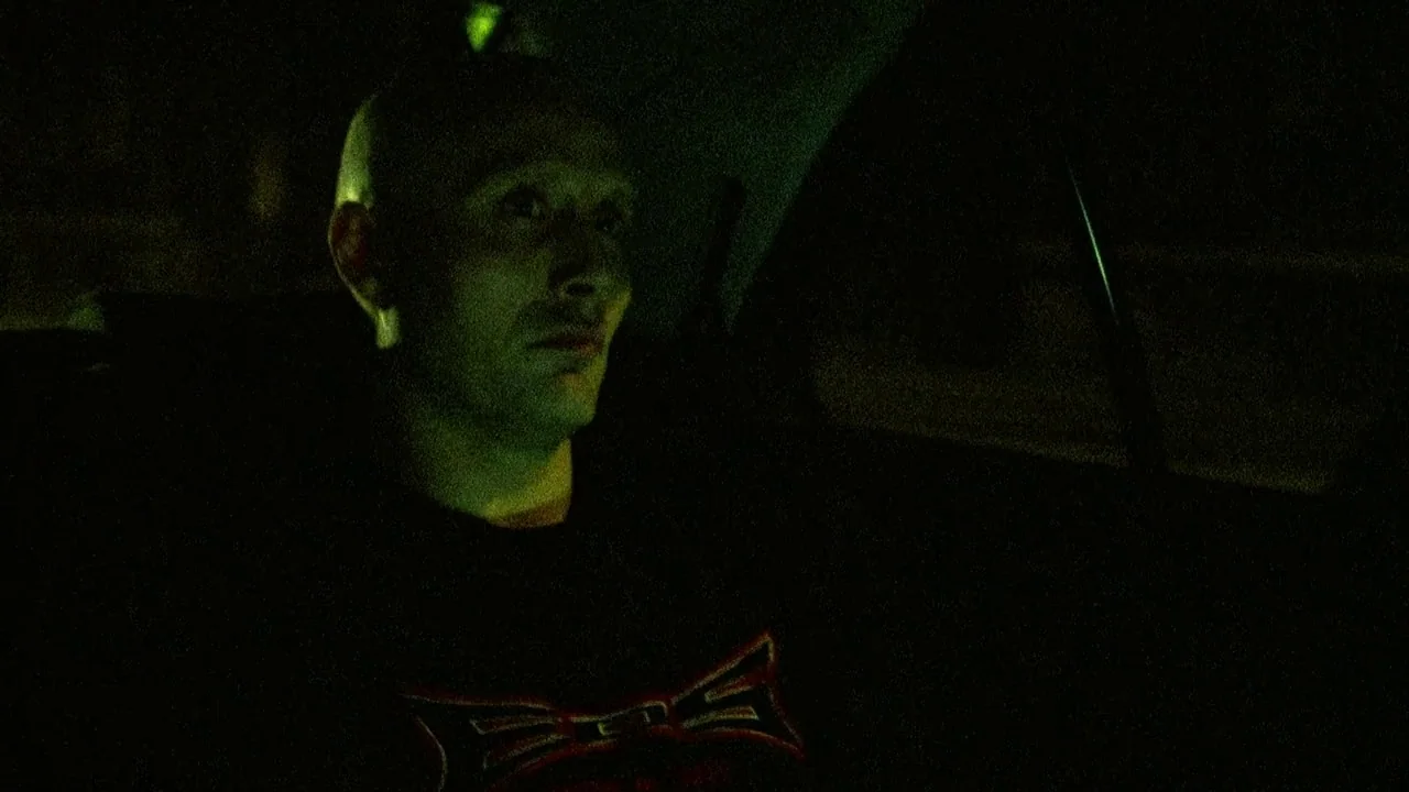 Pusher II (2004) movie still: medium — A man sits inside the darkened cabin of a vehicle at night. Green neon light reflects onto his skin while he…