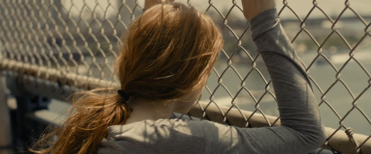 The Disappearance of Eleanor Rigby: Her (2014) movie still: closeup — A person stands on a metal bridge walkway next to a chain-link fence. The…