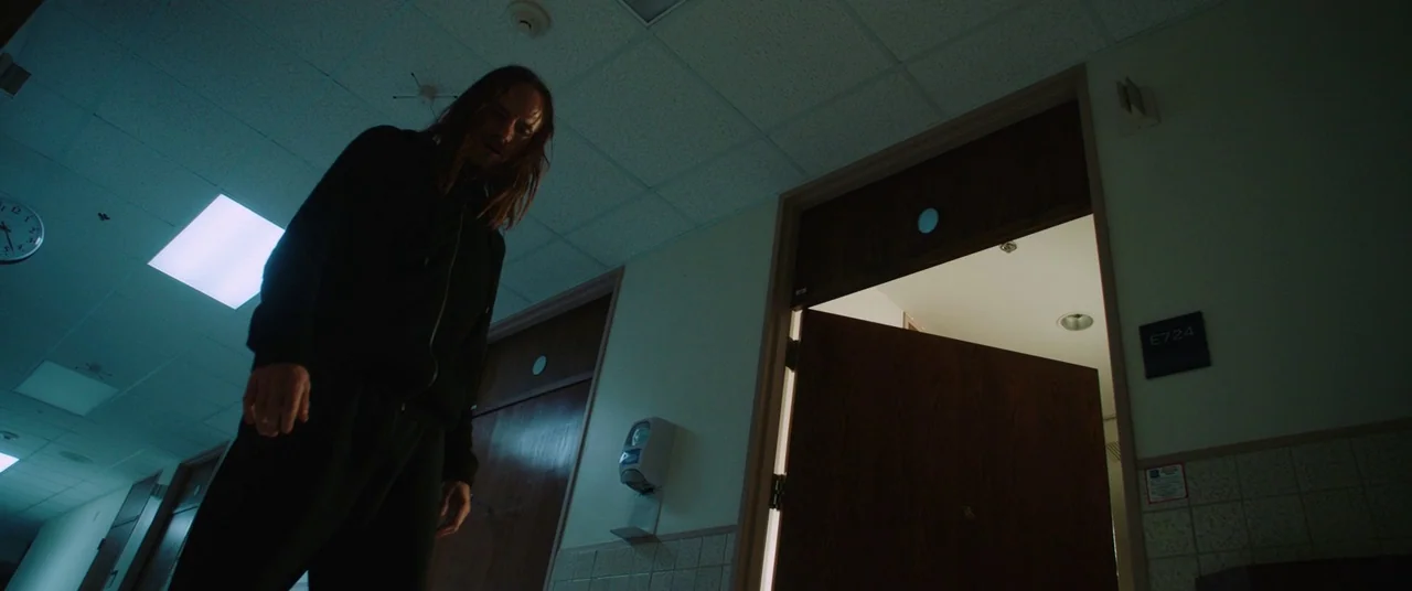 Happy Death Day (2017) movie still: medium — A dimly lit hospital corridor features tan walls and wood-paneled doors with signage. A man with long…