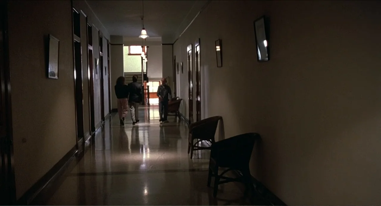Rain Man (1988) movie still: wide — A long, dimly lit indoor hotel corridor has dark walls, framed art, and wicker chairs. Three people walk away…