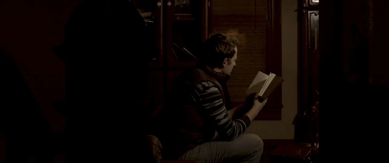 Amityville Cult (2021) movie still: medium — A dark, dimly lit room contains a person reading in a chair. A man in a vest sits holding an open book…