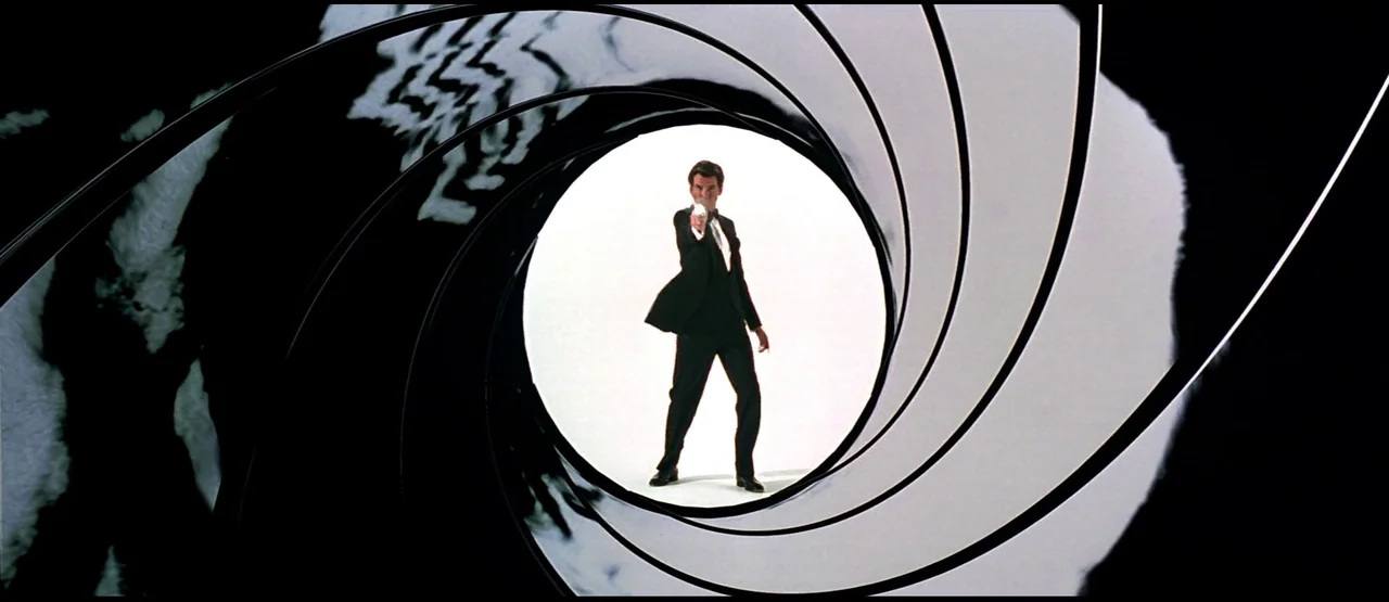 GoldenEye (1995) movie still: fullbody — A man in a formal tuxedo stands centered within a swirling circular graphic. He aims a handgun directly at…