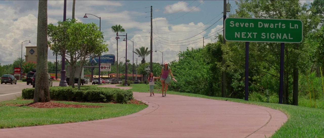 The Florida Project (2017) movie still: wide — A pink paved path curves through a grassy roadside area under a cloudy sky. Two females walk towards…