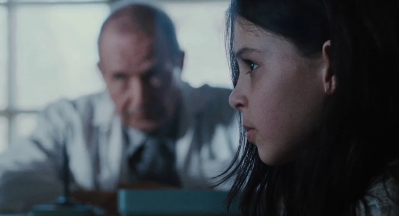 Martyrs (2008) movie still: closeup — A stark office environment features an older man sitting at a desk behind a girl. The girl stands in the…