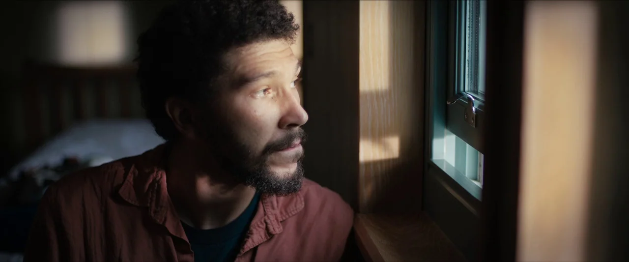In The Earth (2021) movie still: closeup — A man stands indoors next to a wood-framed window during the daytime. The man looks outside while soft…