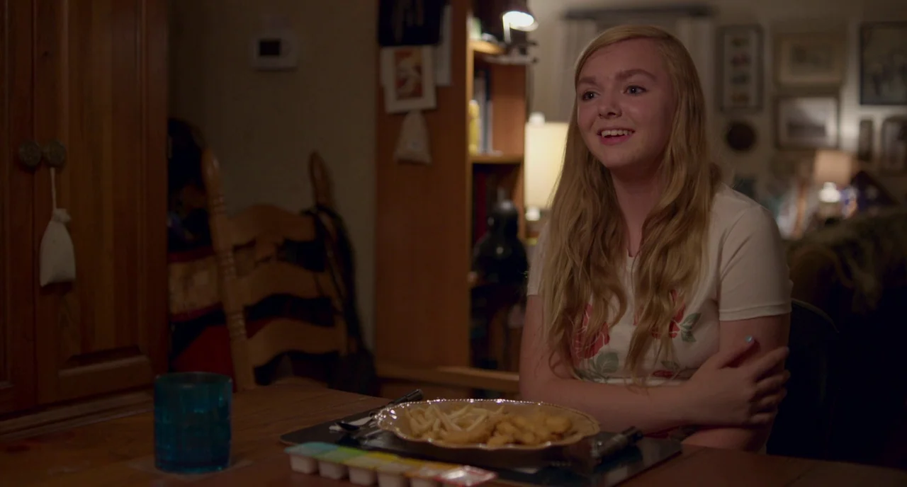 Eighth Grade (2018) movie still: medium — A dimly lit room contains a wooden table with a plate of food. A young woman with blonde hair sits at the…