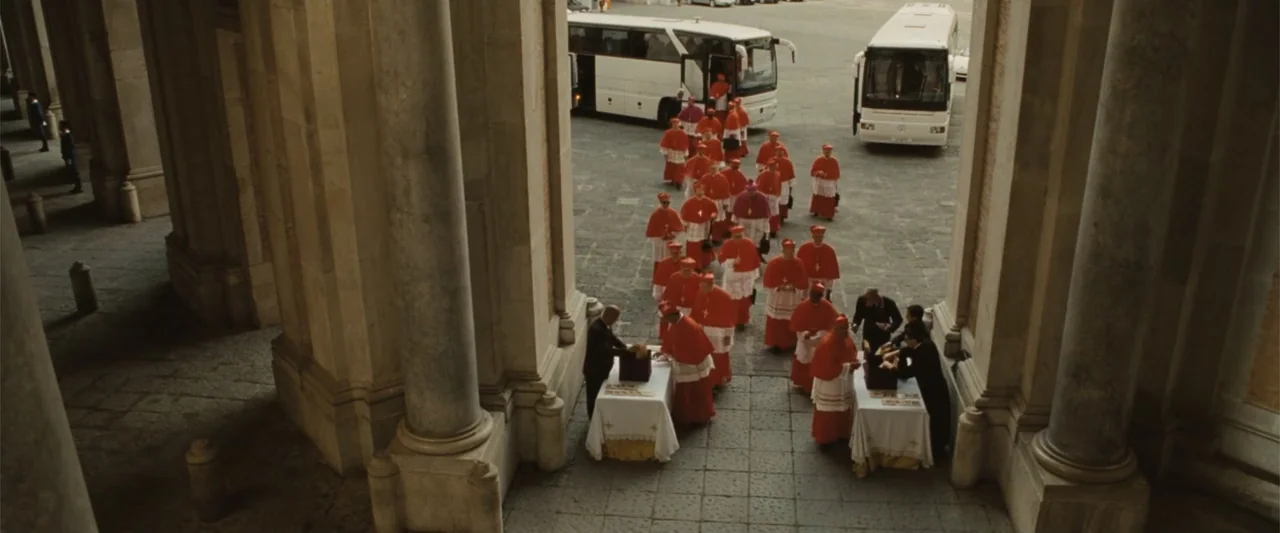 Angels & Demons (2009) movie still: wide — A stone-paved courtyard is flanked by massive stone pillars in a colonnade. A large group of men in red…