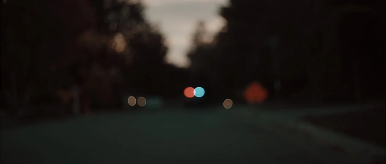 Night of the Missing (2023) movie still: establishing — A dark, out-of-focus suburban road stretches into the dim evening distance. Distant, blurred…