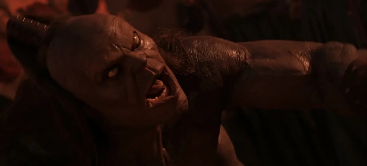 Mortal Kombat (2021) movie still: closeup — A dark subterranean chamber is illuminated by flickering warm firelight. A heavily muscled orc creature…