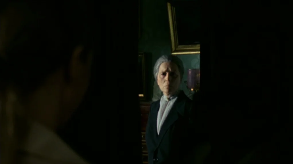 Face to Face (1976) movie still: medium — A dark hallway with green walls and framed portraits dimly lit by a lamp. An elderly woman stands at the…