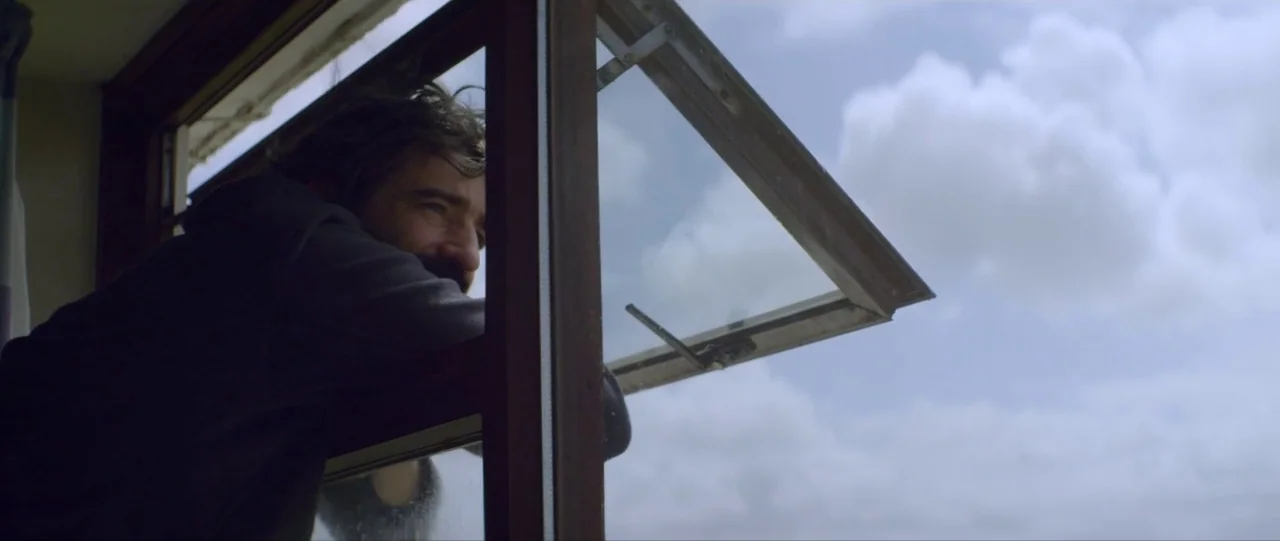Silence (2012) movie still: medium — A man looks through an open window frame towards a bright, cloudy daylight sky. The wooden window frame bisects…