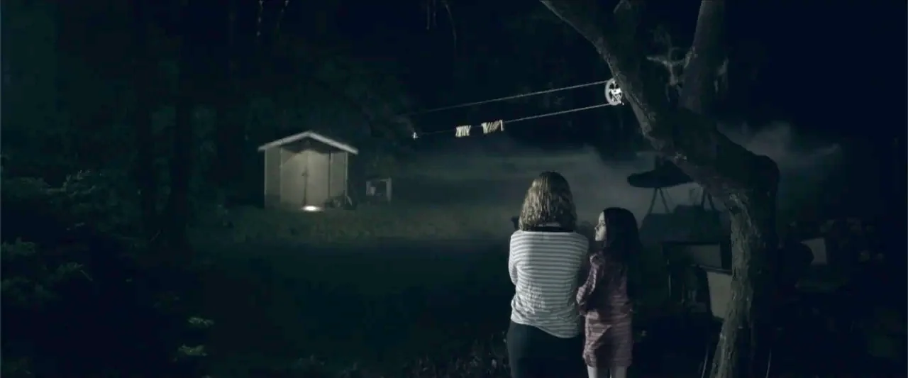 The Hollow Child (2018) movie still: wide — A dark, grassy backyard at night featuring a small shed with a glowing light. Two female figures stand…