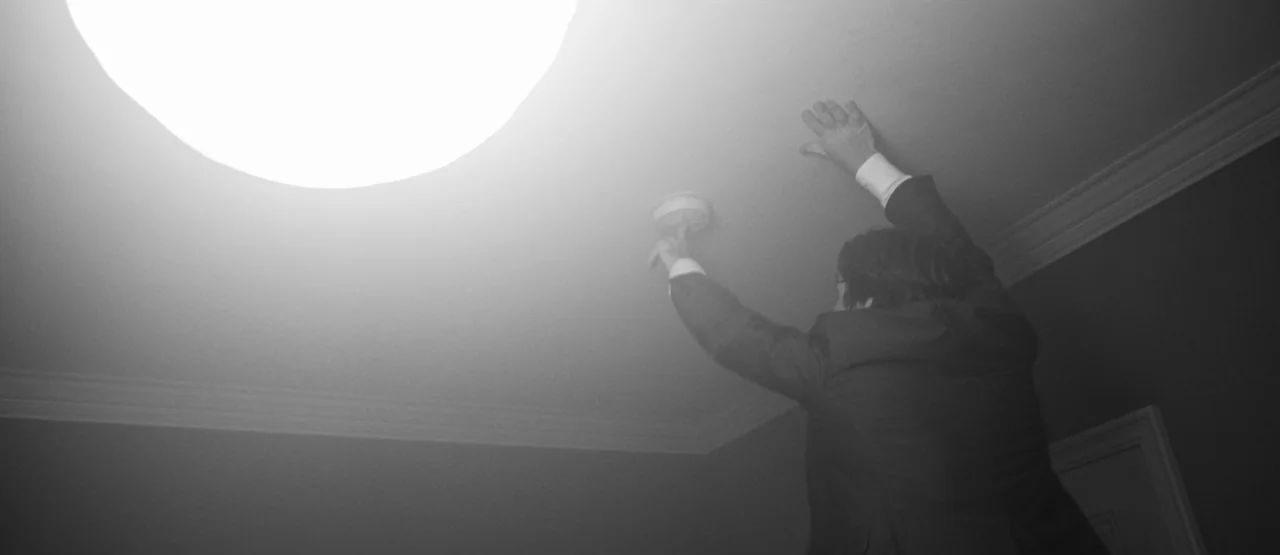 The Party (2017) movie still: medium — A man in a suit reaches up toward the ceiling. He touches a smoke detector while standing near a bright light.