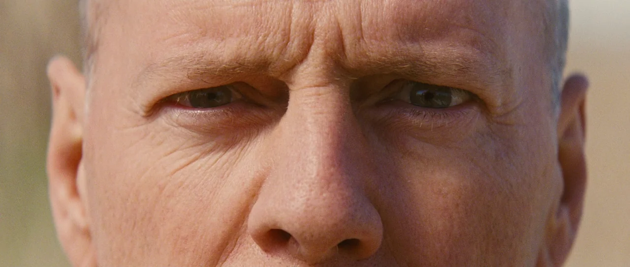 Looper (2012) movie still: closeup — An outdoor, sunlit environment serves as the background for a tight facial portrait. A middle-aged man with…