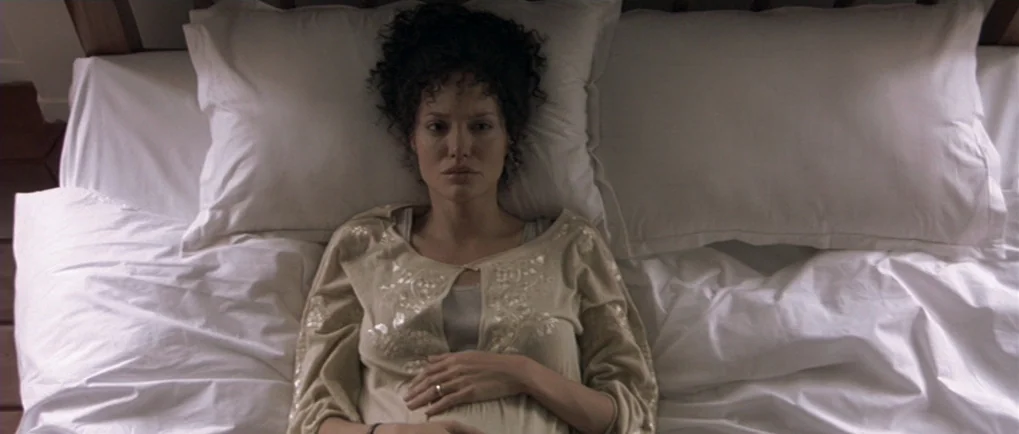 A Mighty Heart (2007) movie still: medium — A woman lies flat on a crisp white bed with large pillows. She stares upward while resting her hands…
