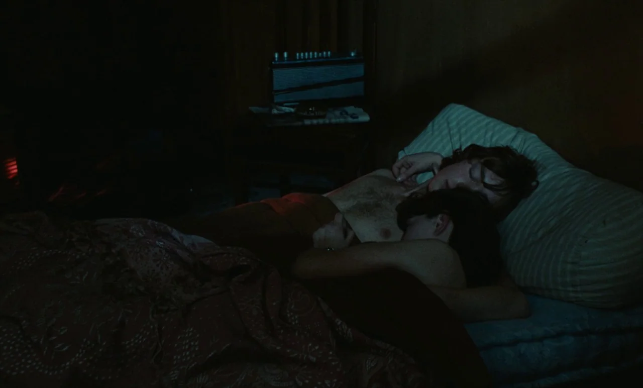 Graduate First (1978) movie still: medium — A dark, cramped room featuring a bed with a patterned quilt and striped pillows. Two figures lie…