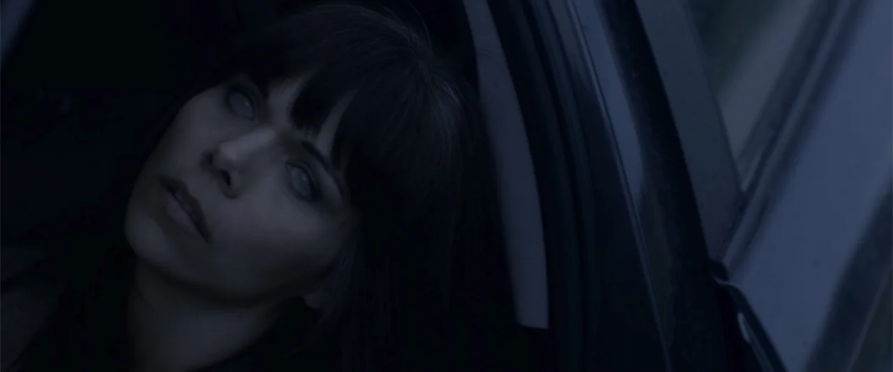 Radius (2017) movie still: closeup — The interior of a car at night featuring a dark cabin and window frame. A woman with dark hair and white eyes…