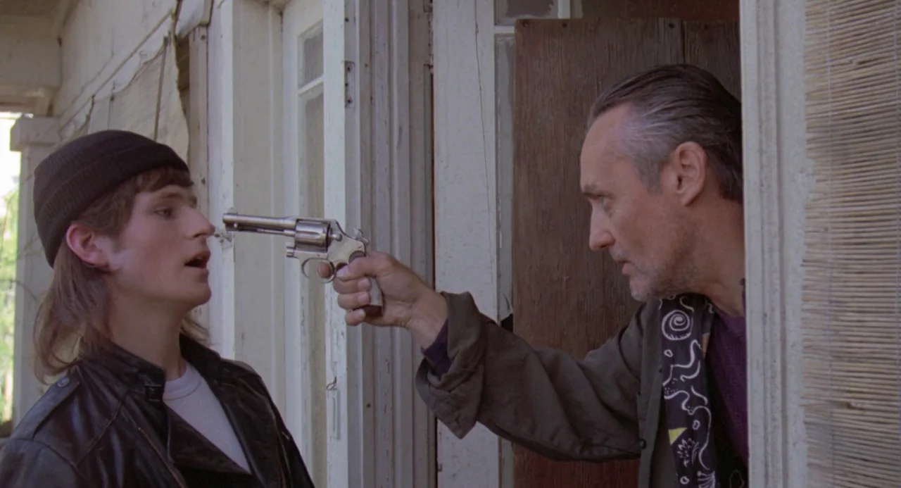River’s Edge (1986) movie still: medium — A wooden porch entrance with peeling paint and a textured bamboo screen. An older man aims a metal…