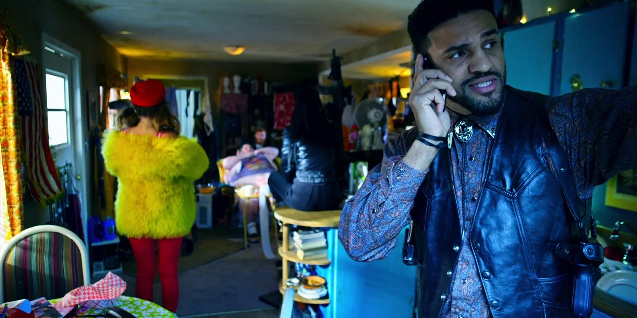 Polar (2019) movie still: medium — A cluttered residential space filled with various household objects and figures in the background. A man stands…