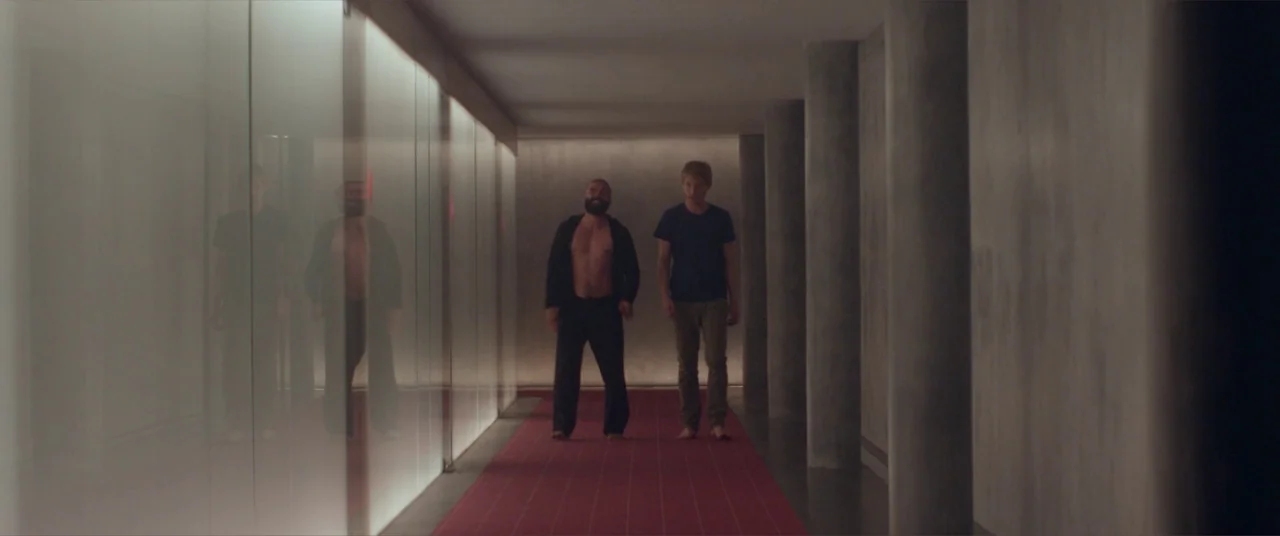 Ex Machina (2015) movie still: fullbody — A long minimalist interior space features polished gray columns and a red carpet runner. Two men walk…