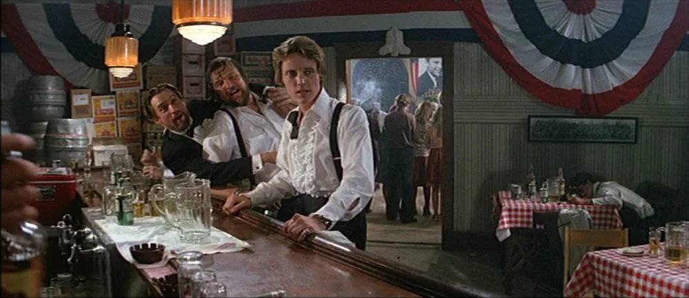 The Deer Hunter (1978) movie still: medium — A wooden bar setting is decorated with red, white, and blue patriotic buntings. Two men lean on the bar…