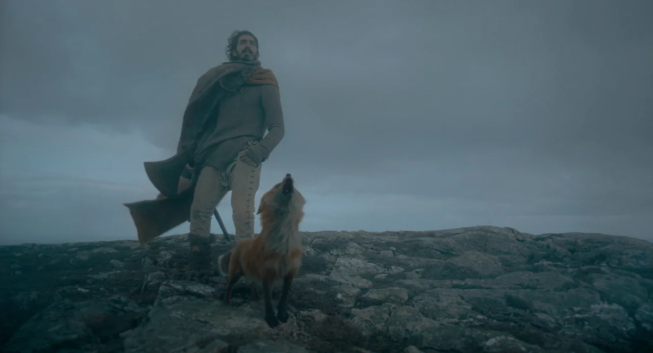 The Green Knight (2021) movie still: fullbody — A man and a fox stand together atop a desolate, rocky, overcast mountain cliff. The man looks toward…