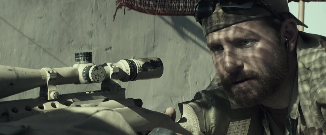 American Sniper (2014) movie still: closeup — A man in combat gear rests against a light-colored stucco wall in bright sunlight. He peers through…
