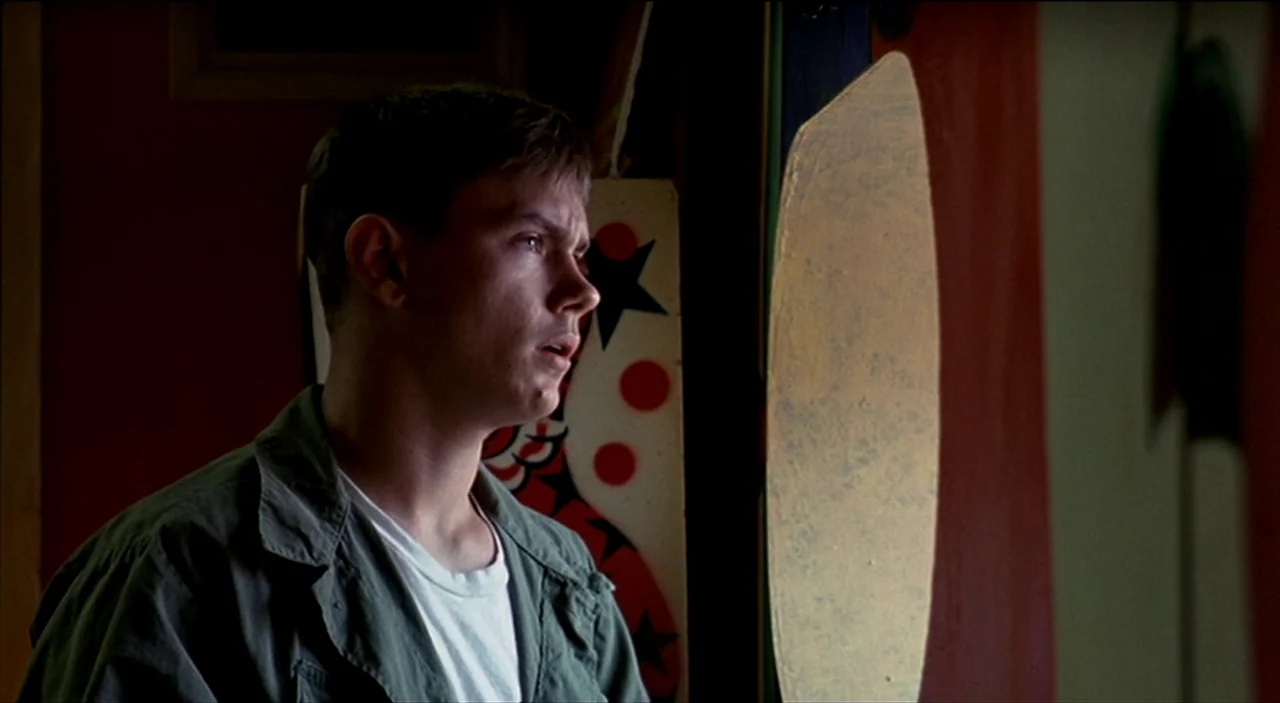 Dogfight (1991) movie still: medium — A dimly lit space features a red and white patterned canvas backdrop. A young man stands in profile looking…