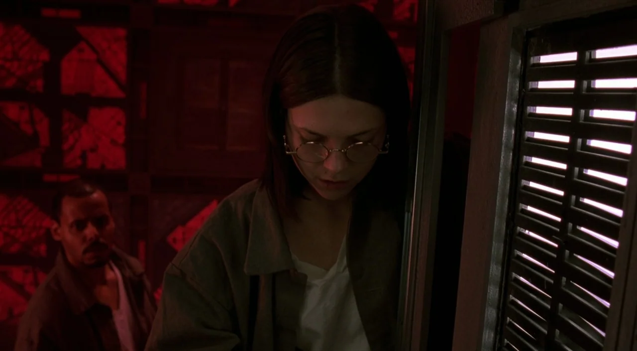 Cube (1997) movie still: medium — An interior space with walls composed of modular dark panels and red neon grids. A woman looks downward while…