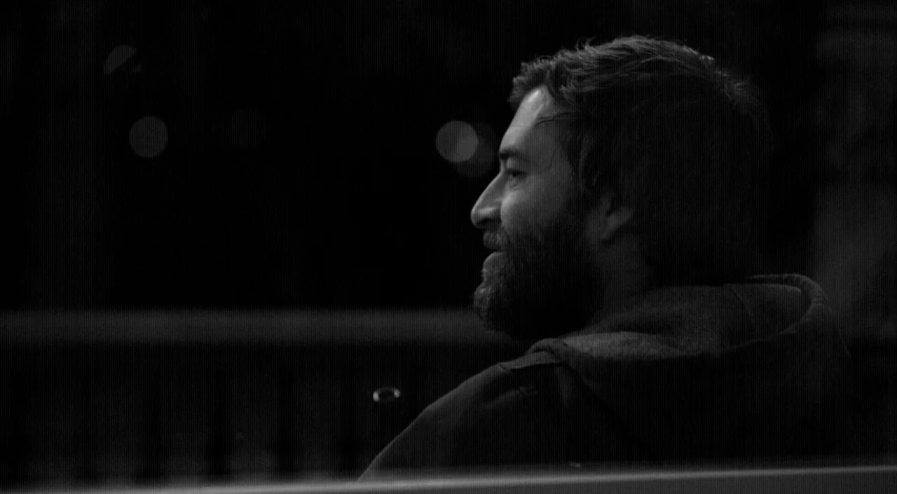 Blue Jay (2016) movie still: closeup — A man sits on a bench at night in a dark outdoor space. He looks toward the side while illuminated by faint…
