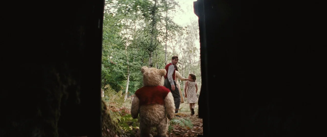 Christopher Robin (2018) movie still: fullbody — A dark, silhouetted foreground doorway opens into a bright, lush wooded area. A man and a girl walk…