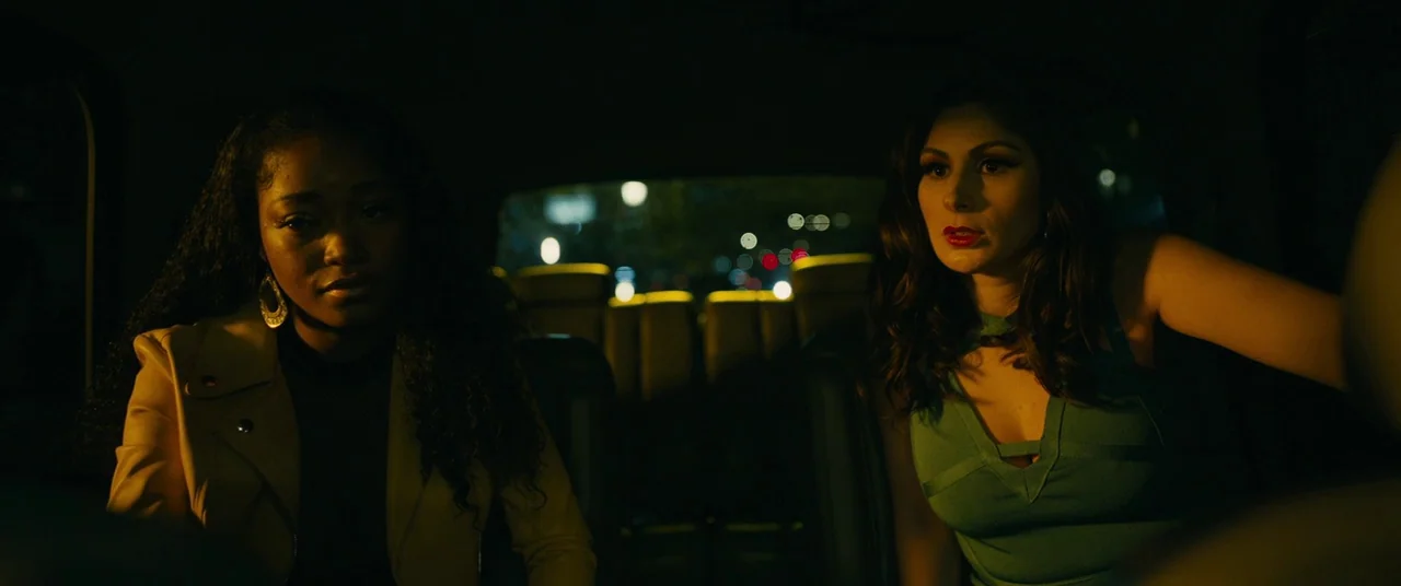 Hustlers (2019) movie still: medium — The interior of a car at night with out-of-focus city lights visible through windows. Two young women sit…