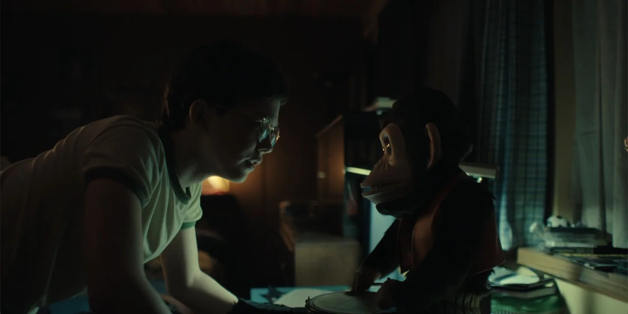 The Monkey (2025) movie still: medium — A dimly lit child's bedroom features wooden furniture and a blue curtain background. A young boy in glasses…